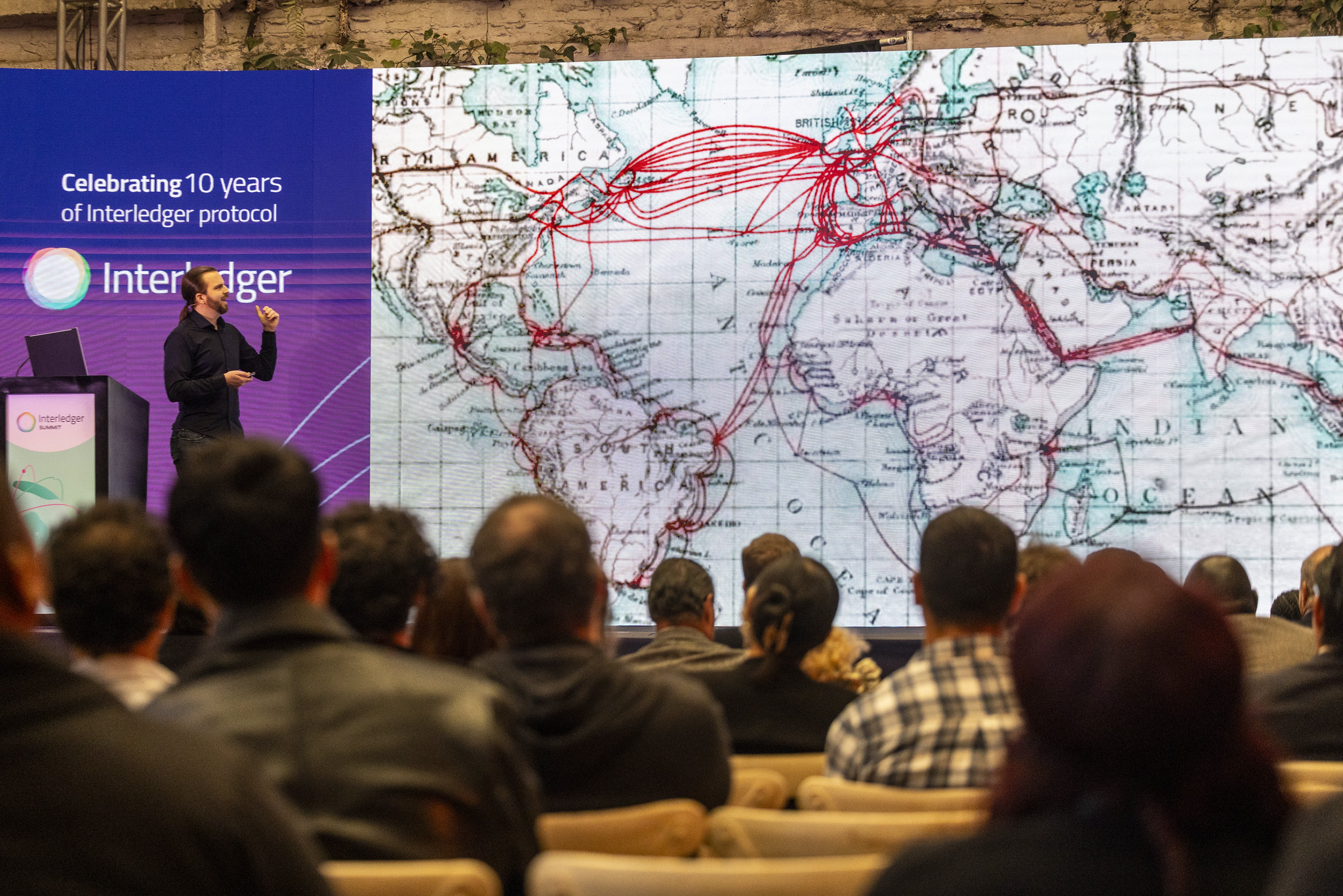 A speaker presenting at the Interledger Summit beside a world map of payment routes.